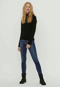 Črn pulover z dolgimi rokavi in visokim ovratnikom, kombiniran s tesno prilegajočimi modrimi kavbojkami iz jeansa ter črnimi gležnjarji, na svetlem ozadju.