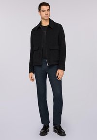 Chaqueta de mezcla de lana negra con cuello, dos bolsillos frontales, combinada con jeans de mezclilla azul oscuro y zapatos de cuero negros.