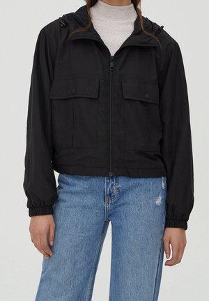 Person iført sort lynlåstrøje med lommer med klap foran, elastiske manchetter, over en grå rullekrave, kombineret med lyseblå, revnede jeans.