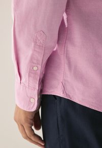 Camicia rosa chiaro con finitura testurizzata, dotata di polsini con bottoni e dettagli alle cuciture laterali, abbinata a pantaloni blu scuro.