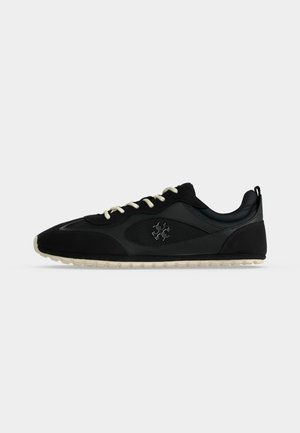 Zwarte lage sneaker met witte veters, beige zool, mesh- en lederen details en een klein metalen logo aan de zijkant.