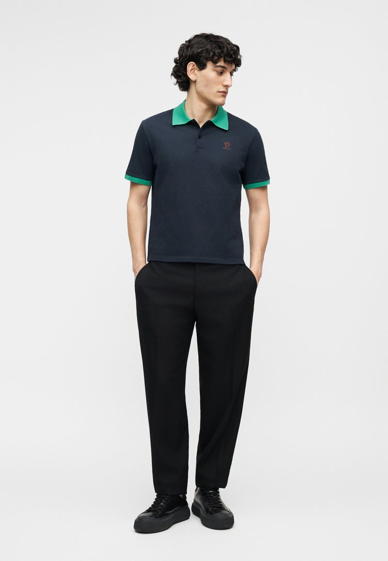 Man die een marineblauw poloshirt met groene kraag en manchetten draagt, zwarte broek en zwarte sneakers, staand met handen in de zakken en zijwaarts kijkend.