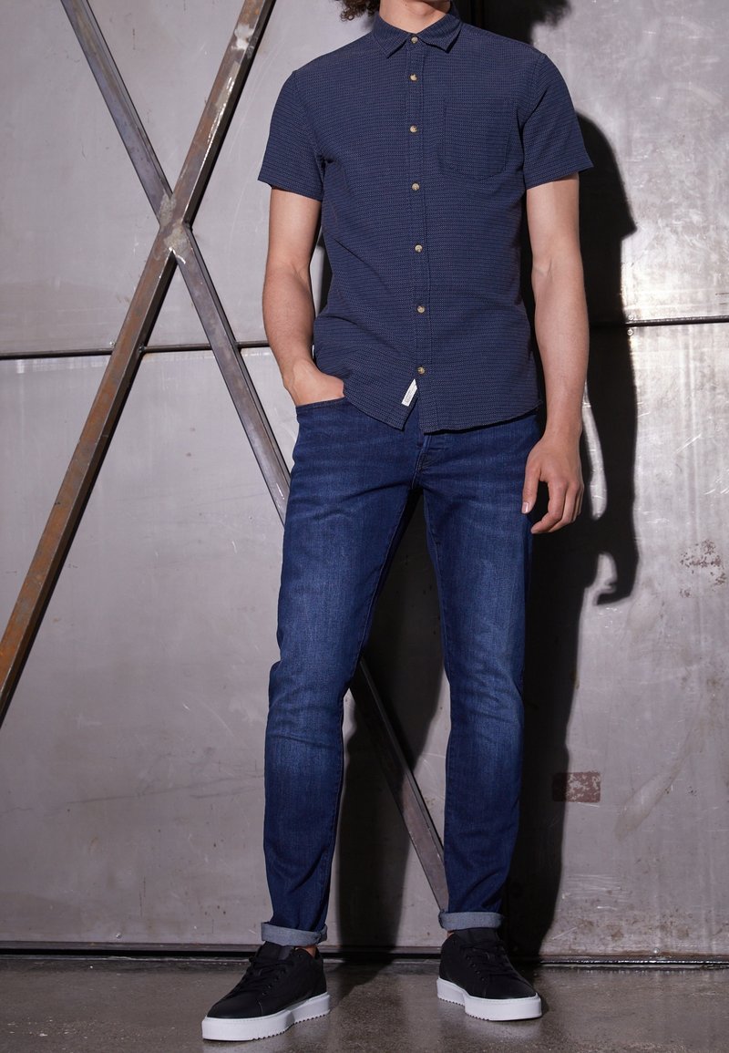 Homme portant une chemise bleu marine à manches courtes boutonnée, un jean bleu foncé retroussé et des baskets noires, debout avec une main dans la poche.