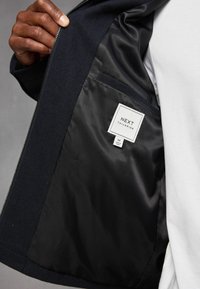 Giacca nera con un interno liscio e lucido. Presenta un'etichetta di "NEXT TAILORING" all'interno di un quadrato bianco e una tasca interna visibile.