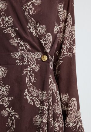 Bruine jurk met witte bloemen- en paisleypatronen, textuurstof en een gouden decoratieve knop op de taille.