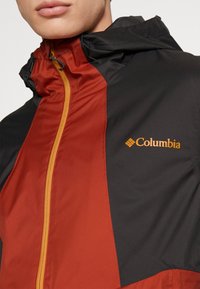 Homme portant une veste Columbia noire et orange rouille avec une fermeture éclair jaune et un logo sur la poitrine, partiellement fermée jusqu'au cou.