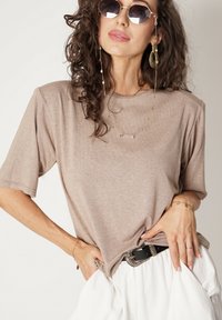 Beżowa koszulka z krótkim rękawem o luźnym kroju, wykonana z miękkiego materiału. Charakteryzuje się okrągłym dekoltem i jest zestawiona z czarnym paskiem oraz białymi spodniami.