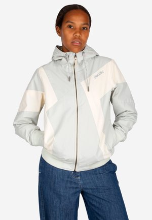 Frau trägt eine hellgraue und weiße Kapuzenjacke mit Reißverschluss, die Hände in den Taschen, und blaue Jeans, sie schaut nach vorne.