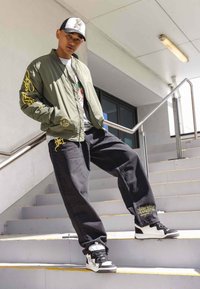 Chaqueta bomber verde con texto amarillo, camiseta gráfica, jeans negros holgados y zapatillas negras y blancas en una escalera con bordes amarillos.