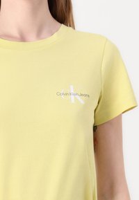 Svetložlté bavlnené tričko s krátkymi rukávmi. Na ľavej strane hrudníka sa nachádza biele potlačené logo "Calvin Klein Jeans". Mäkká textúra.