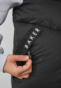 Crni prsluk s punjenjem i kockastom teksturom, s velikom oznakom "BAKER" na bočnom džepu i rukavom svijetlosive boje u pozadini.