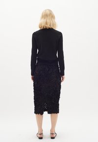 Top nero a maniche lunghe abbinato a una gonna a tubino nera con texture e dettagli floreali a strati. Il modello indossa sandali neri con fasce.