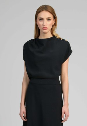 Femme blonde portant une robe noire sans manches avec des détails à boutons sur l'épaule, debout devant un fond gris uni.