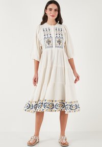 White cotton dress with embroidered floral patterns in blue and yellow. Features puff sleeves and a tiered skirt design.