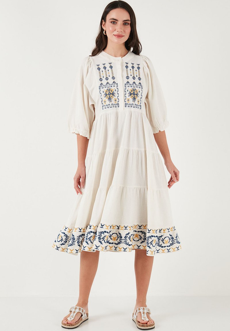 White cotton dress with embroidered floral patterns in blue and yellow. Features puff sleeves and a tiered skirt design.