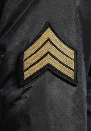 Schwarzer Stoff mit einem goldenen, gestickten Chevron-Abzeichen, das drei nach oben gerichtete Streifen zeigt, die von schwarzen Nähten umrandet sind.