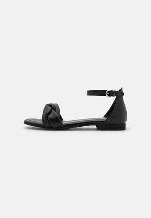 Sort flad sandal med knyttet forrem og ankelrem med sølvspænde på ensfarvet baggrund.