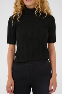 Žena oblečená v čiernom rebrovanom krátkom roláku a čiernych nohaviciach s vysokým pásom pred jednoduchým pozadím.