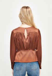 Blouse en satin marron à manches longues, avec un détail froncé dans le dos et une découpe en forme de trou de serrure. Assortie à un jean en denim bleu avec des poches arrières.