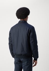 Chaqueta bomber de color azul marino con una textura suave, cuello y puños acanalados. Combinada con jeans de mezclilla oscuros. La vista muestra la parte trasera.