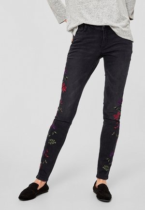 Woman wearing black slim-fit jeans with floral embroidery on legs, paired with black loafers and a light gray sweater.