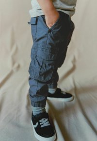 Dunkelblaue Cargo-Hosen mit elastischen Bündchen, ausgestattet mit Seitentaschen. Kombiniert mit schwarzen Turnschuhen und grauen Socken. Glatte Stofftextur.