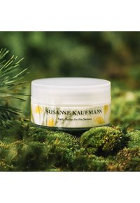 Pot circulaire blanc de beurre corporel avec un couvercle transparent, orné d'illustrations de poissons jaunes et de texte noir sur un fond vert naturel.