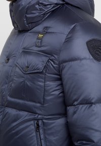 Mörkblå puffjäcka med glänsande yta, utrustad med en stor huva, dragkedje- och tryckknappsfickor samt en patchlogotyp på axeln.