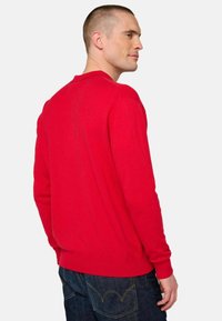 Roter Pullover aus weichem Stoff mit rundem Ausschnitt, gerippten Bündchen und geradem Saum, kombiniert mit dunklen Jeans.