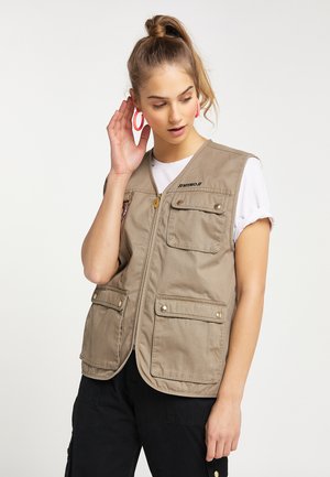 Junge Frau mit hohem Pferdeschwanz, die eine beige Utility-Weste über einem weißen T-Shirt und schwarzen Hosen trägt und mit der linken Hand einen pinken Creolen-Ohrring berührt.