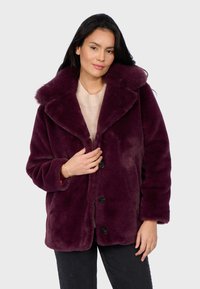 Mujer vestida con un abrigo de pelaje morado profundo sobre un suéter beige, con pantalones negros, de pie contra un fondo gris claro liso.