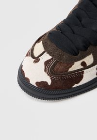 Καφέ δερμάτινο αθλητικό παπούτσι με λεπτομέρειες σε μοτίβο αγελάδας. Διαθέτει μαύρα υφασμάτινα κορδόνια και λαστιχένια σόλα. Στρογγυλερή μύτη και ραμμένες λεπτομέρειες.