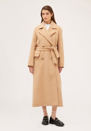 Abrigo beige de doble botonadura con cinturón, que presenta una tela texturada, cuello con muescas y mangas largas, llegando por debajo de las rodillas.