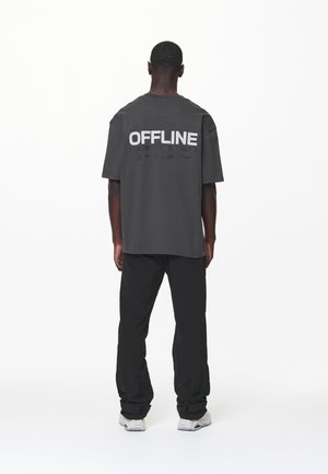 Szara, oversize koszulka z napisem "OFFLINE" w pogrubionym białym tekście na plecach, zestawiona z czarnymi spodniami i jasnymi butami.