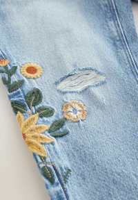 Tissu denim bleu clair avec une déchirure et des fleurs jaunes et orange brodées avec des feuilles vertes le long du bord.