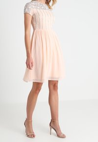 Meruňkov dress s přiléhavým živůtkem, zdobenými rukávy a rozšířenou sukní. Tkanina je lehká s hladkým povrchem.