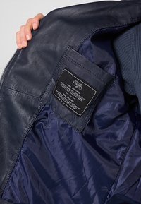 Marinblå läderjacka med en slät nylonfoder inuti. En svart etikett med vit text som anger skötselråd och material.