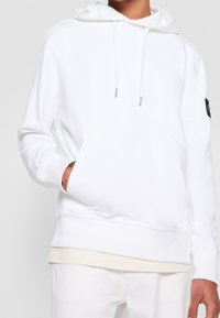 Vit hoodie med framficka, dragsko i huvan, ribbade muddar och ett svart logopatch på ärmen. Mjuk, strukturerad tyg.