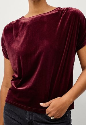 Persoon draagt een kort mouws bordeaux fluwelen top met een ronde hals en donkere spijkerbroek, hand rust in zak met zilveren ring zichtbaar.