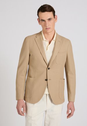 Mężczyzna ubrany w beżową marynarkę na kremowej koszulce polo i białe spodnie, stojący na tle jednolitego, jasnoszarego tła.