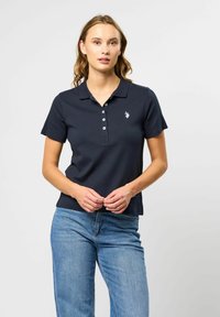 Ung kvinne i mørk navy kortermet poloshirt med knappestolpe og lyseblå jeans, stående foran en ensfarget lys bakgrunn.