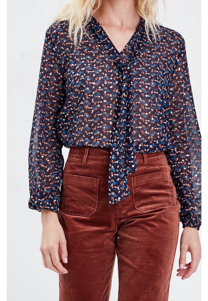 Blouse légère et transparente bleu marine avec un motif de cœurs colorés, dotée d'un col à nouer et de manches longues légèrement bouffantes. Associée à un pantalon en velours côtelé couleur rouille.