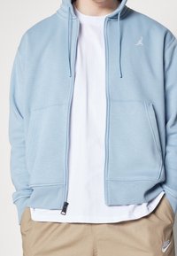 Ljusblå zip-up hoodie i mjukt tyg, med en kenguruficka och en subtil logotyp på vänster bröst. Bärs över en vit t-shirt.