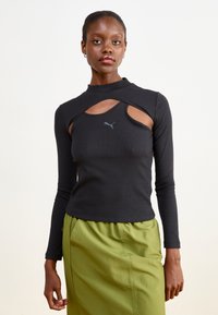 Svart ribbete langermet topp med høy utringning og utskjæringer, med en liten grå Puma-logo på brystet. Kombinert med et grønt skjørt.