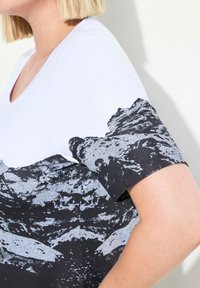 Bílý tričko s černým a stříbrným potiskem hor na krátkých rukávech. Materiál je hladký a má design s výstřihem do V.