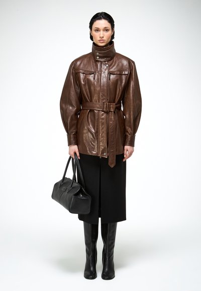 Femme portant une veste en cuir marron ceinturée, une jupe noire et des bottes noires montant jusqu'aux genoux, tenant un sac à main noir, debout devant un fond blanc.