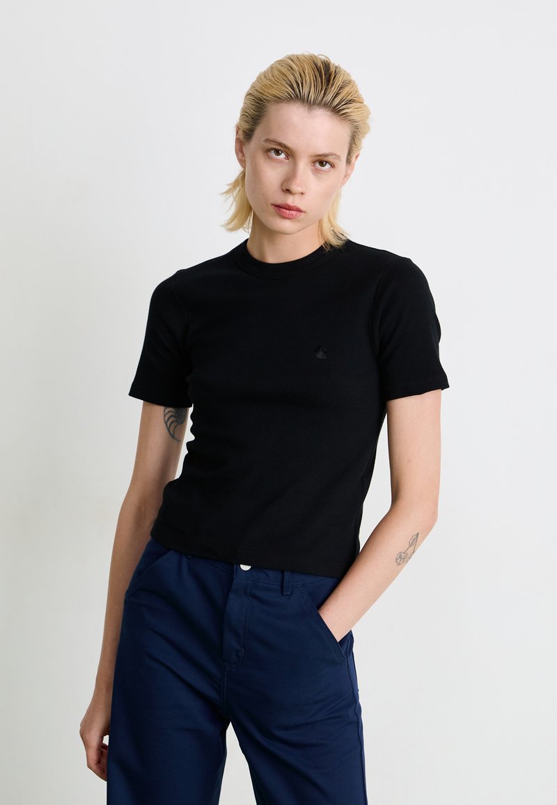 Sort ribbet kortærmet T-shirt med rund hals og lille logo på brystet, båret sammen med marineblå bukser, glat tekstur.