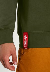 Felpa verde con polsini e orlo a coste, con un'etichetta rossa in tessuto con la scritta "RIMUOVERE PRIMA", parzialmente visibile su pantaloni beige.