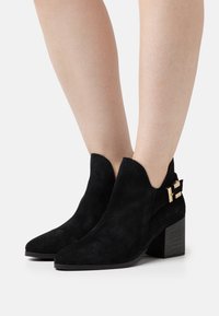 Schwarze Wildleder-Stiefeletten mit einer spitzen Zehenpartie, gezacktem oberen Rand und einem goldenen Schnallenakzent an der Seite. Blockabsatz mit einer strukturierten Oberfläche.