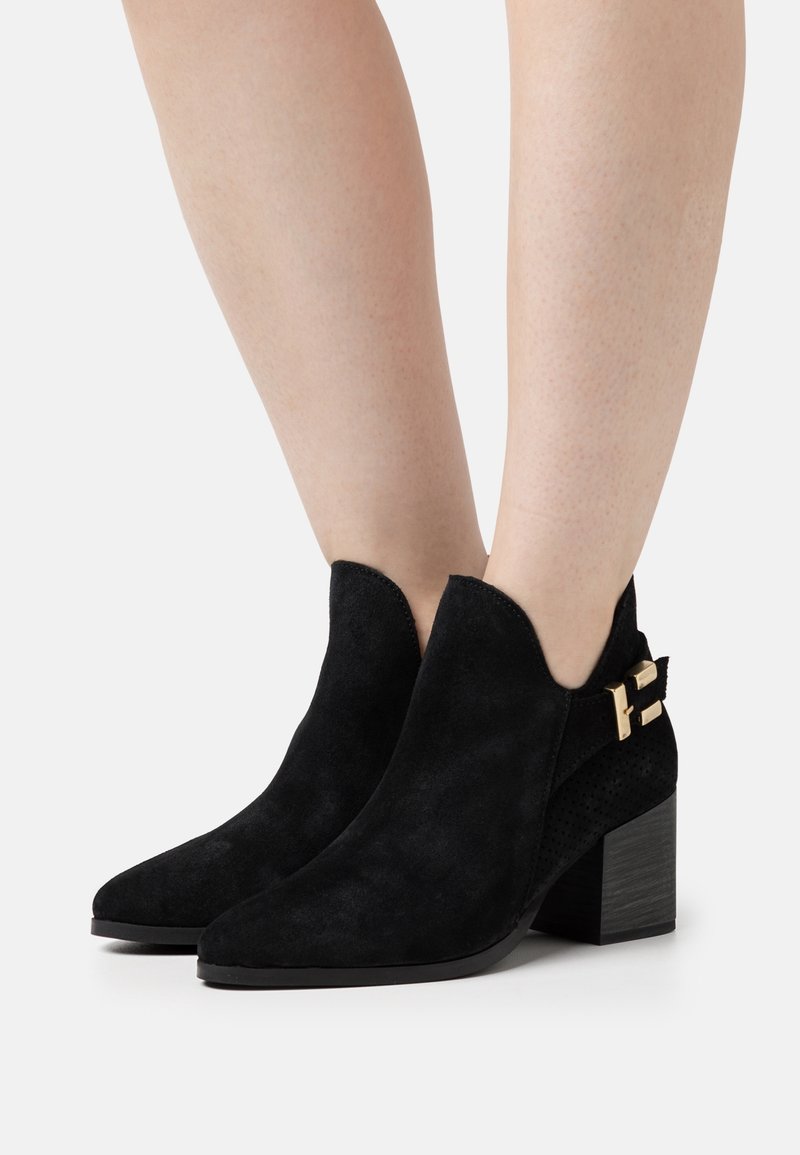 Schwarze Wildleder-Stiefeletten mit einer spitzen Zehenpartie, gezacktem oberen Rand und einem goldenen Schnallenakzent an der Seite. Blockabsatz mit einer strukturierten Oberfläche.